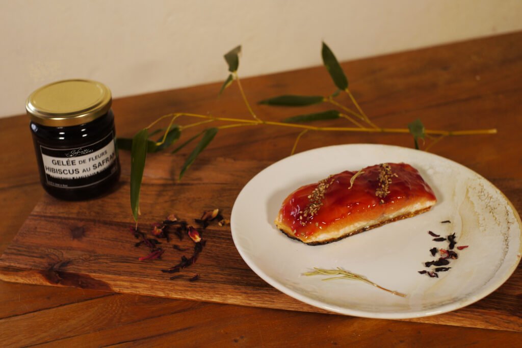 Sur une assiette posée sur une planche de présentation, un pavé de saumon recouvert degelée de fleurs d'Hibiscus au safran prêt à être dégusté. En arrière plan à gauche, un pot de gelée de fleurs d'Hibiscus au safran